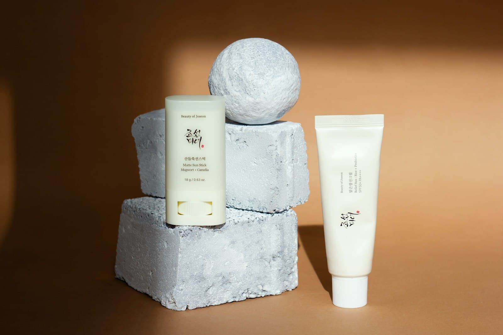 A sunscreen stick and a cream tube displayed on stone blocks against a warm brown background