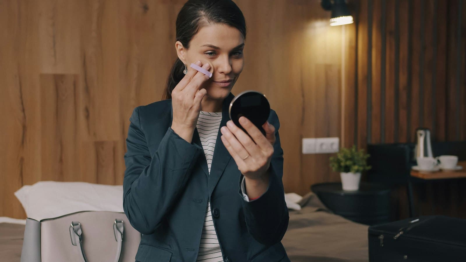 Woman in a blazer pressing a powder puff to her cheek while checking a compact mirror