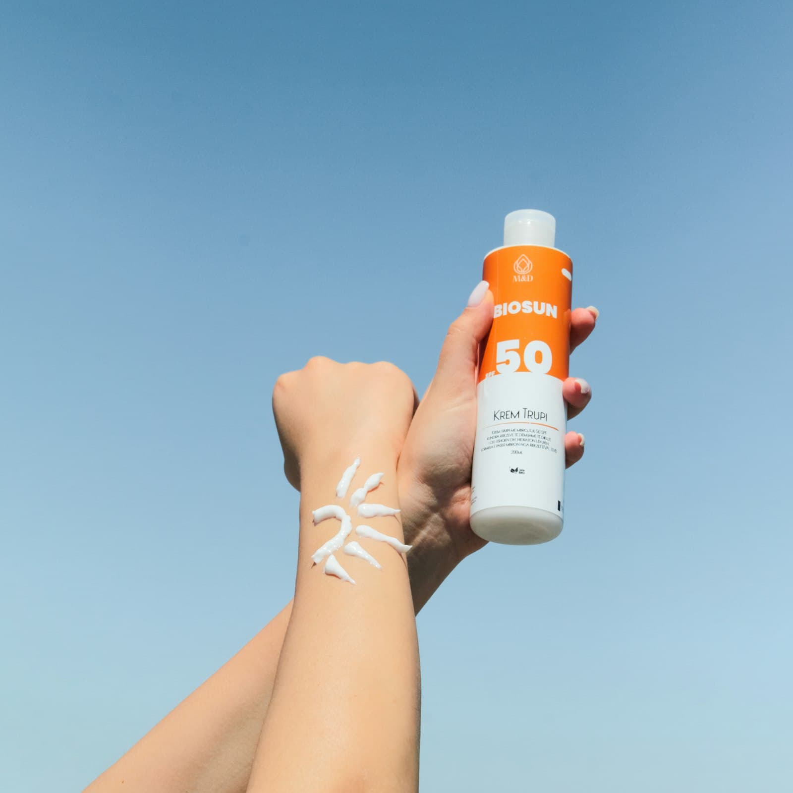 Hand holding an SPF 50 sunscreen bottle beside a sun shape drawn in sunscreen on the wrist, against a blue sky