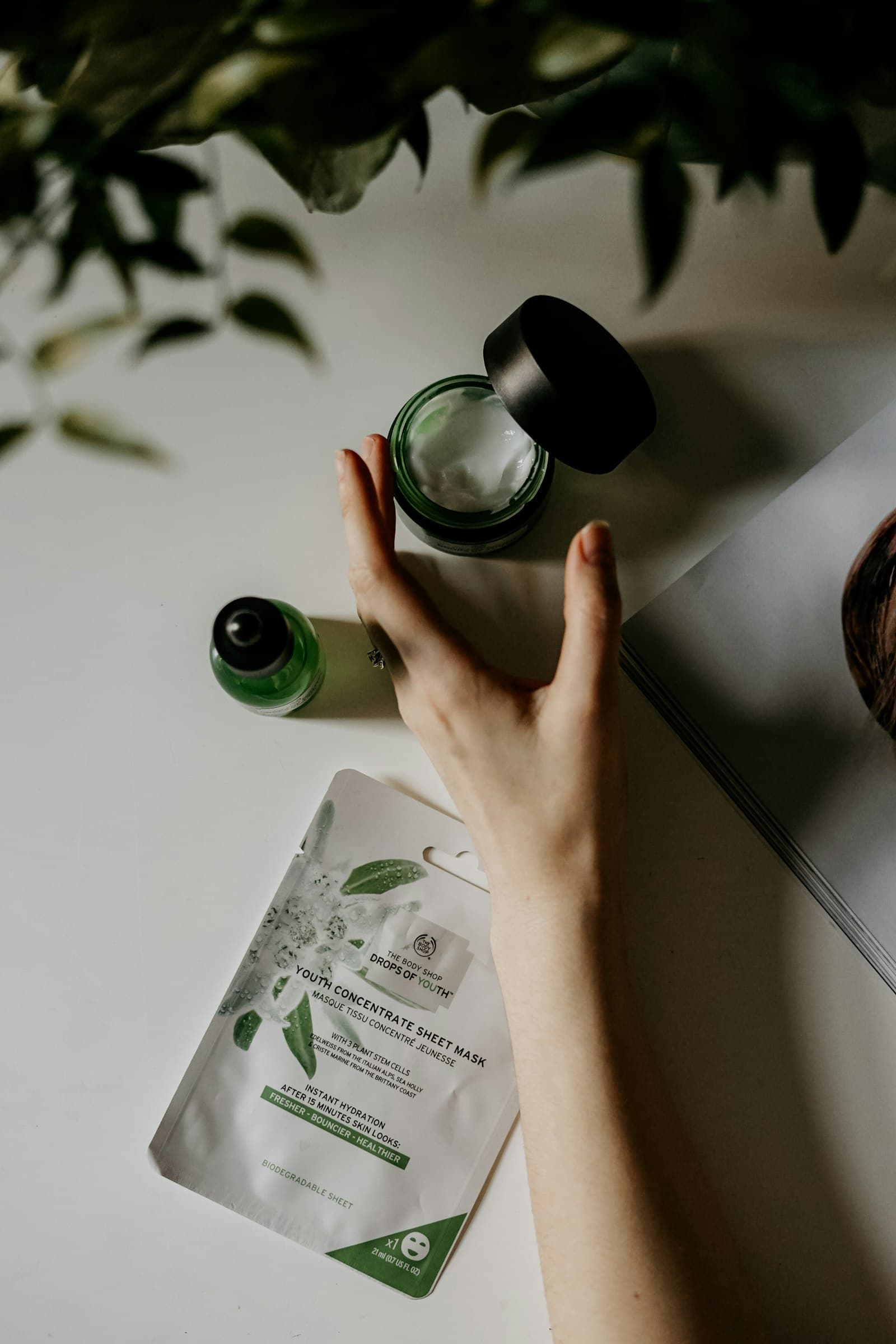 Hand reaching for a jar of cream beside a serum bottle and a sheet mask on a white surface