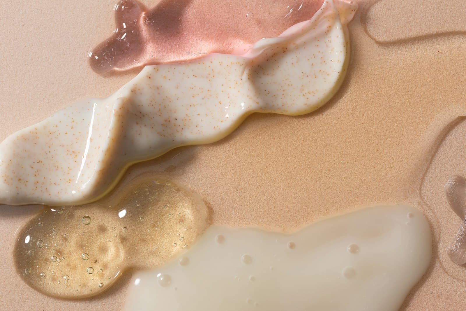 Swatches of a thick white cream, a pink gel and a clear serum spread side by side on a beige surface