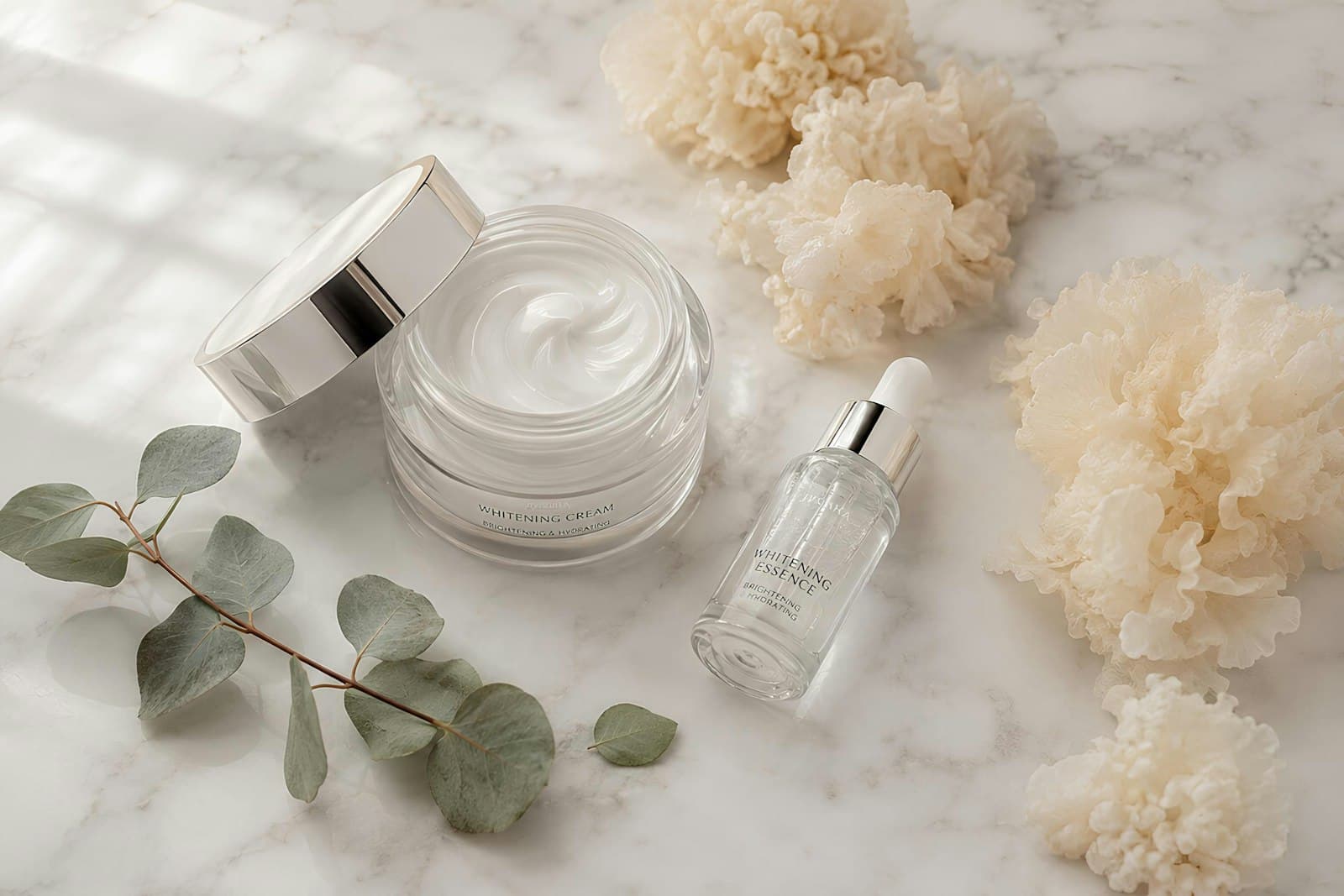Open jar of white face cream beside a serum bottle, eucalyptus and dried flowers on marble