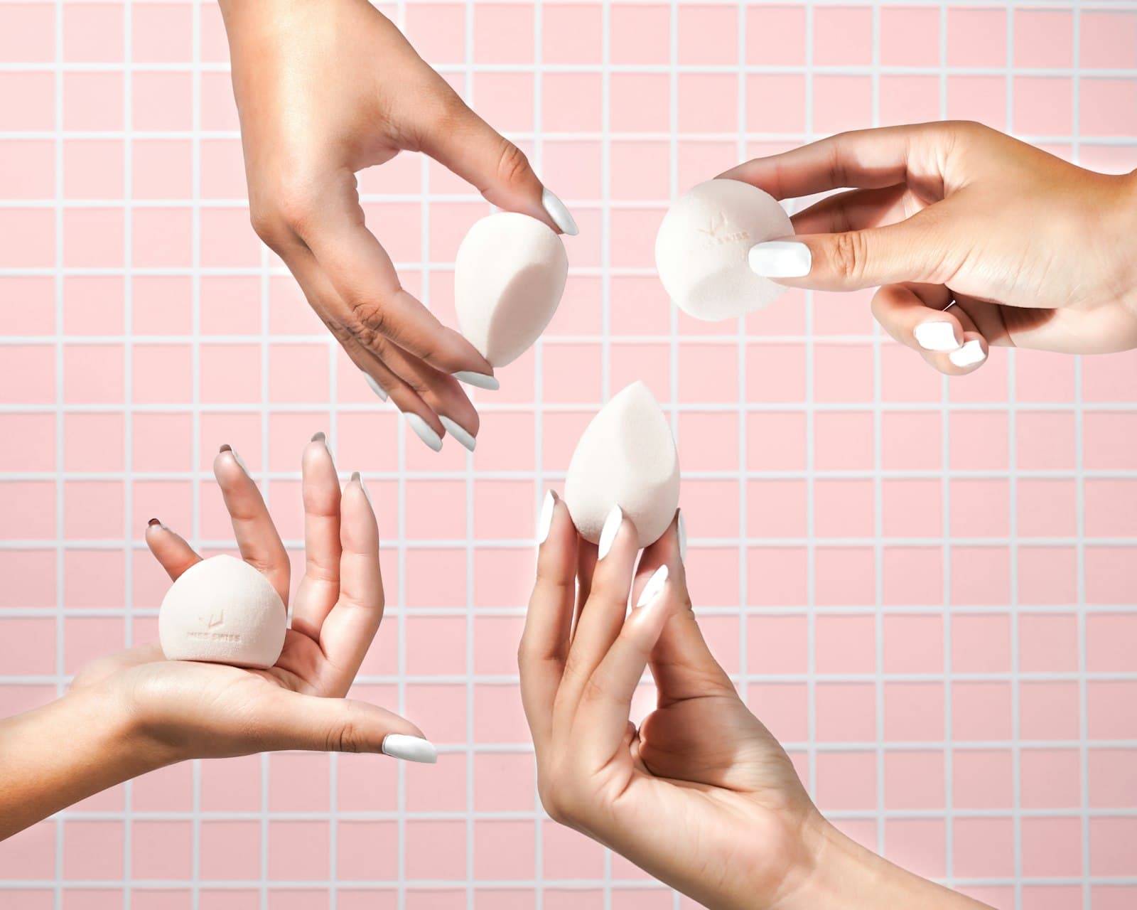 Four hands holding white makeup sponges against a pink tiled background
