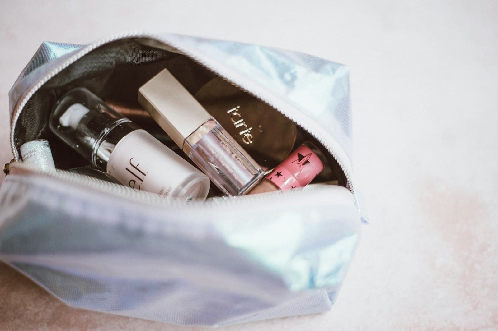 Open iridescent makeup bag holding a foundation bottle, a lip gloss and other small makeup products