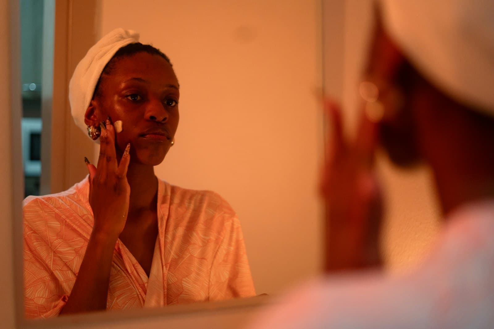 Woman with a towel on her head applying skincare in a mirror in warm evening light