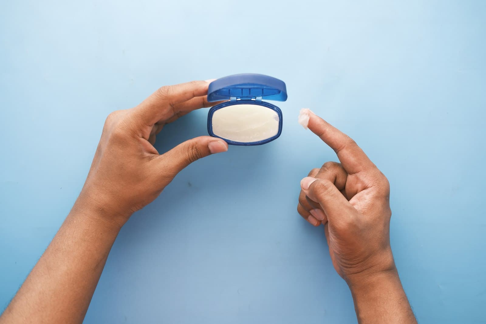Hands holding an open tub of petroleum jelly, with a small dab on one fingertip