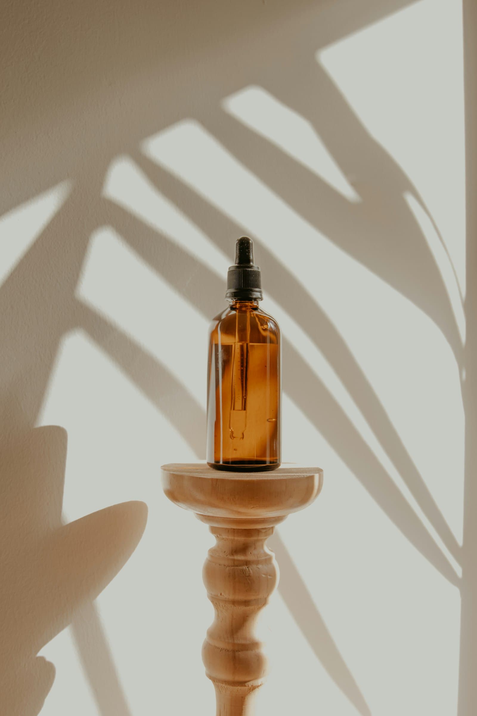 Amber glass dropper bottle on a wooden pedestal with palm leaf shadows on a light wall