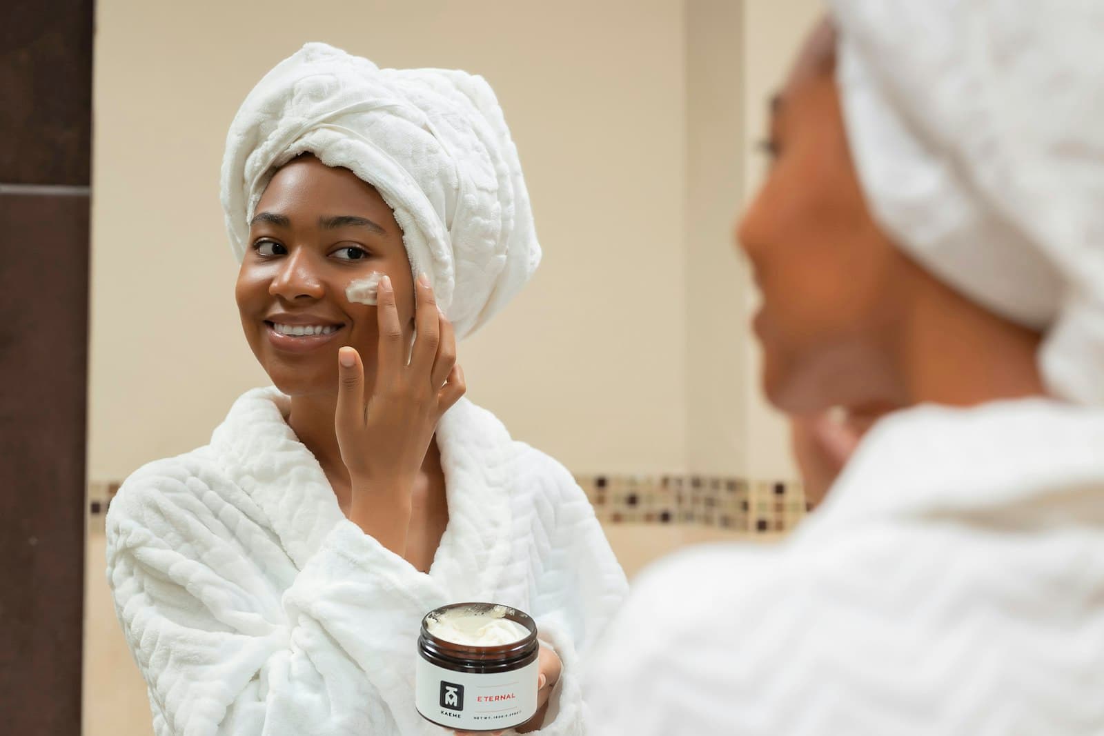 Smiling woman in a towel and robe applying cream from a jar to her cheek in a bathroom mirror