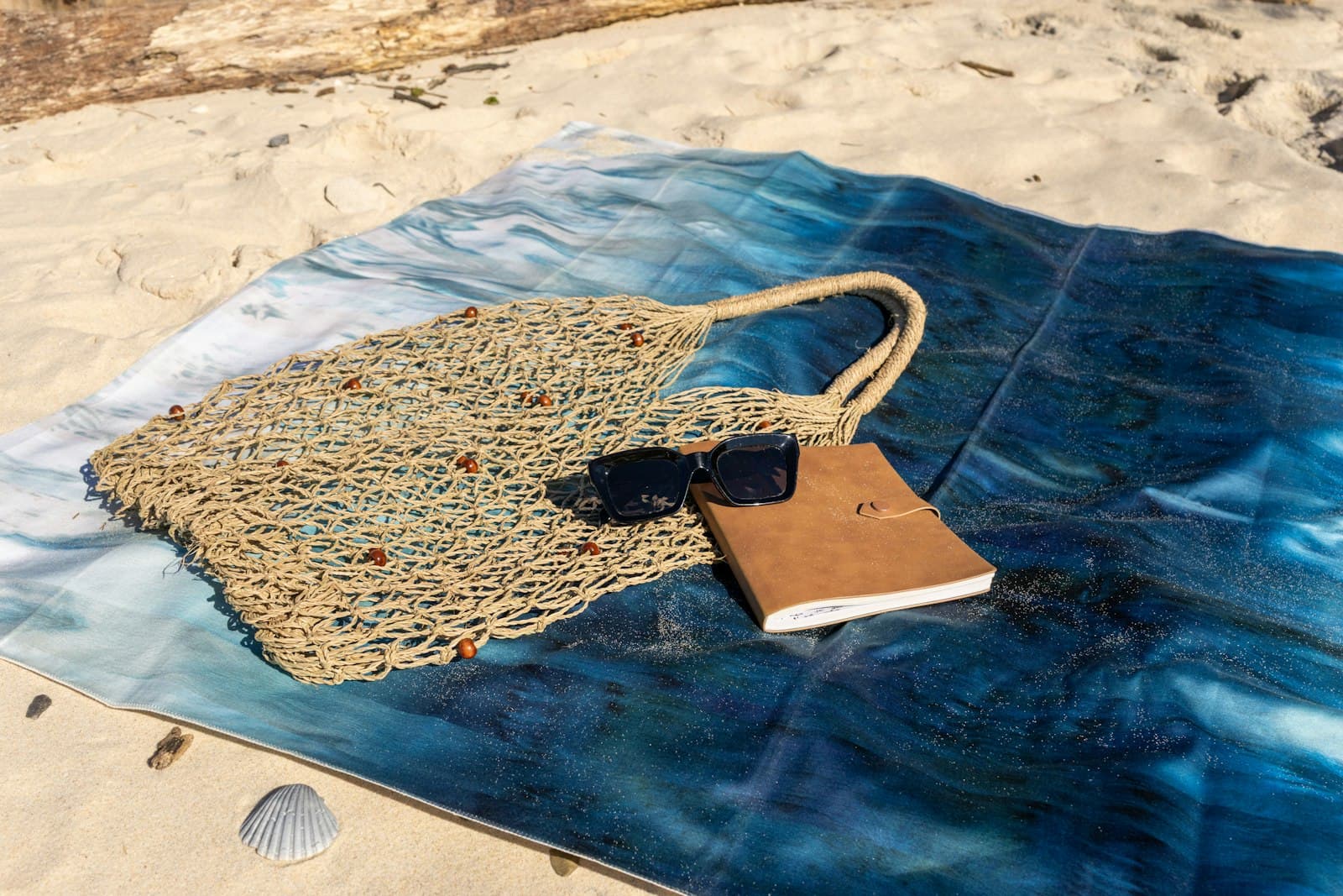 Woven beach bag, sunglasses and a book on a blue towel spread over the sand