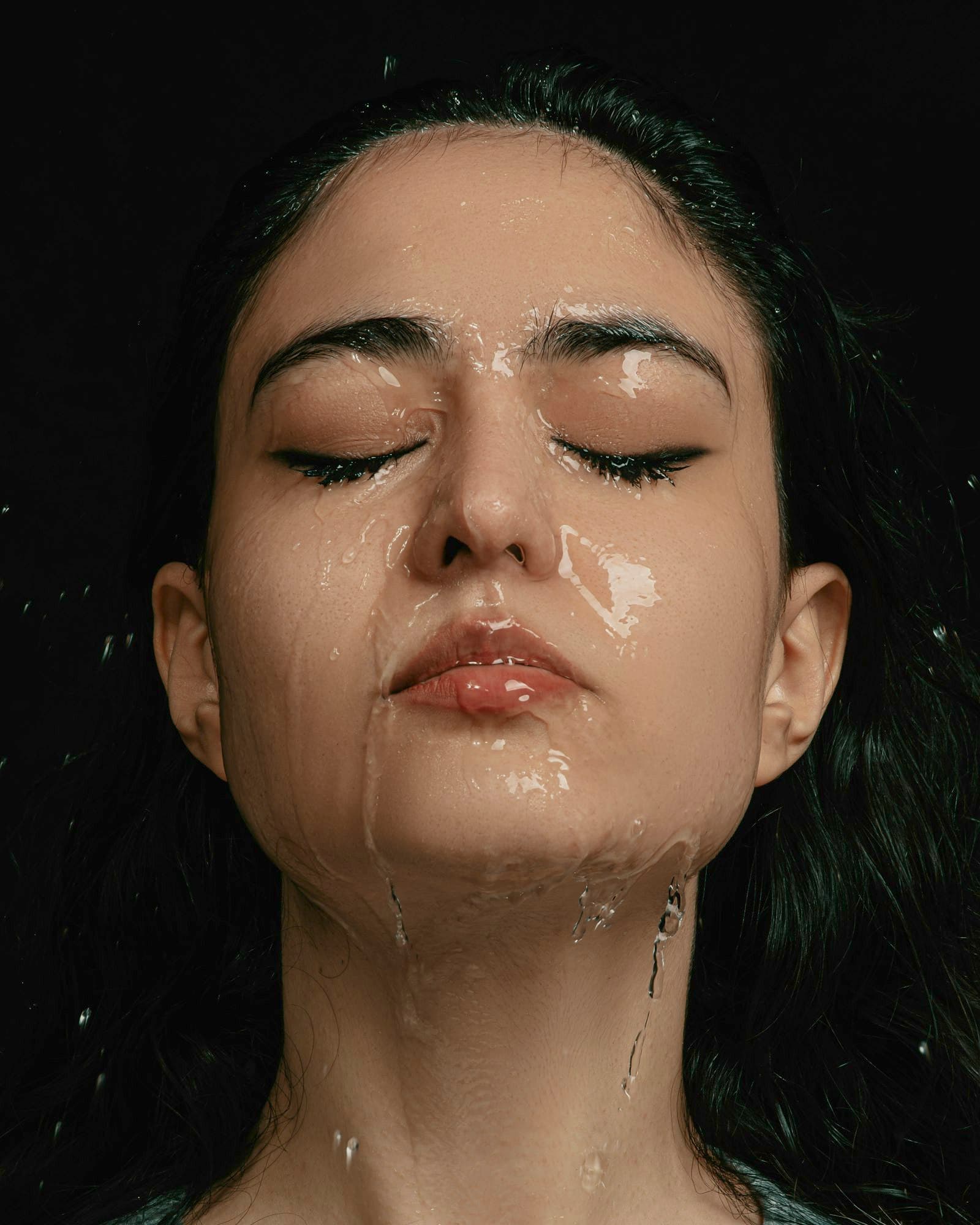Woman with eyes closed and water running down her face against a dark background
