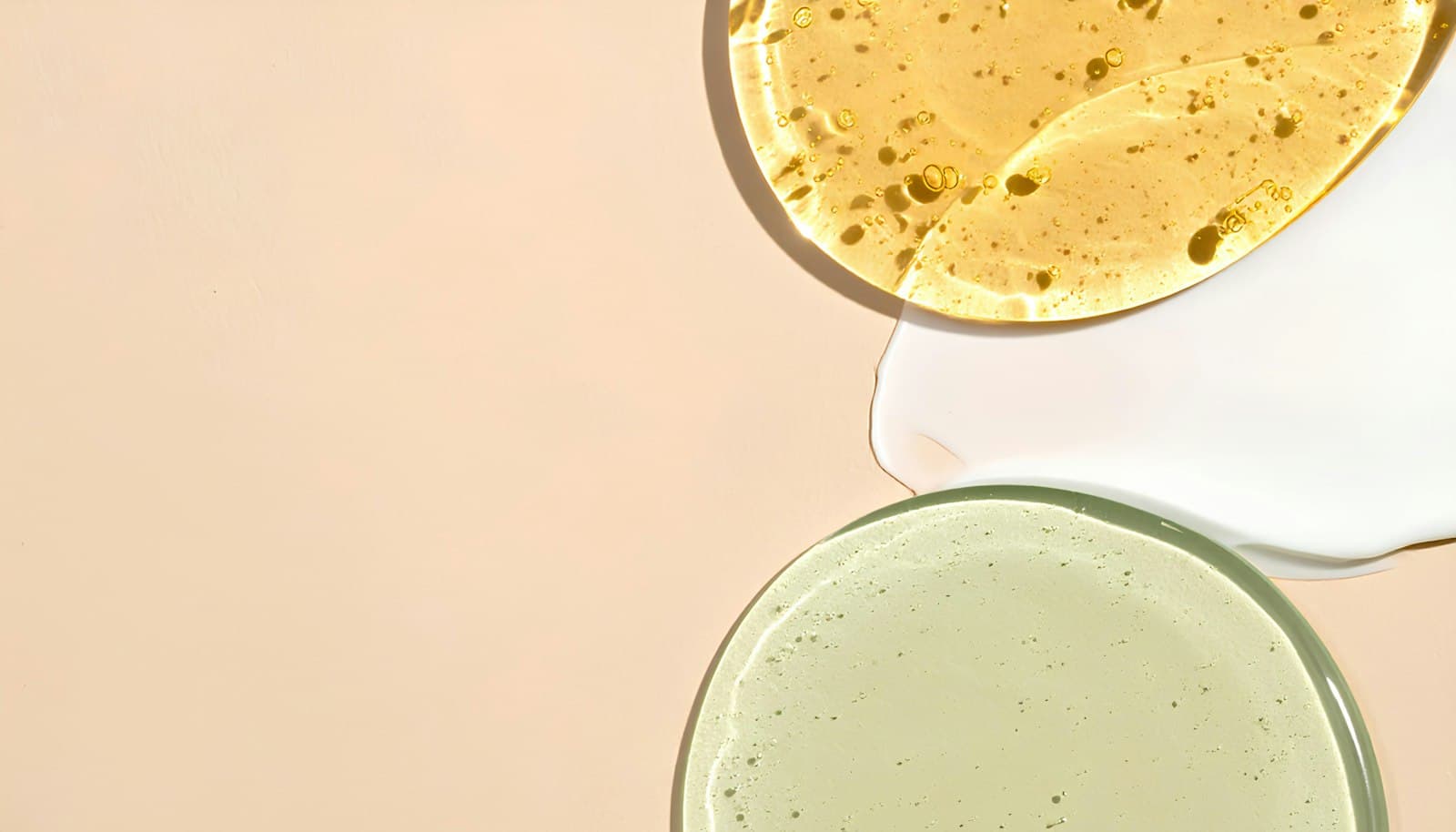 Swatches of a golden serum, a white cream and a pale green gel on a peach background